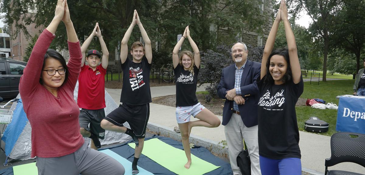 A group of students on yoga matts with their arms above their heads standing on one foot