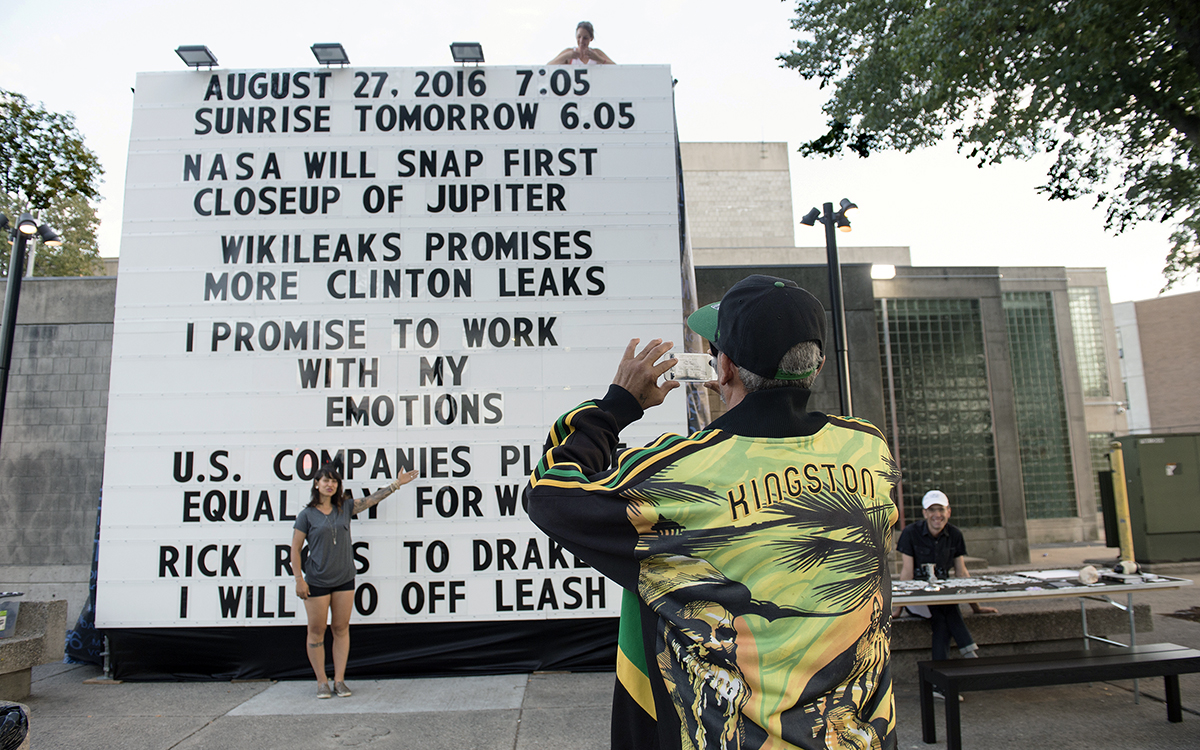 a large white sign with black text in public space with people interacting