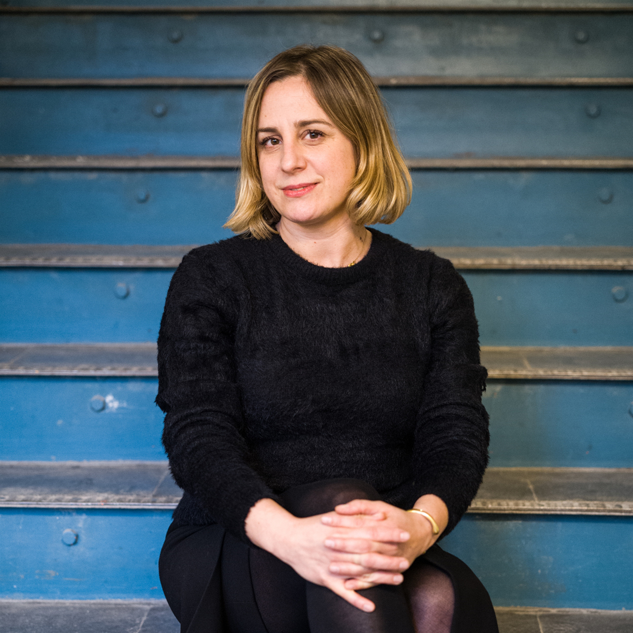 A woman in a black sweater is seated with a blue backdrop.