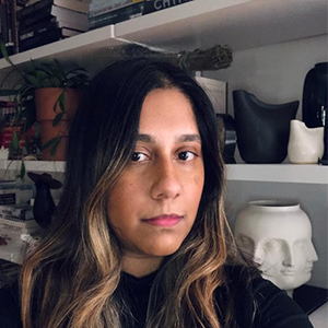 A person with highlighted dark brown hair standing in front of shelves with sculptures.