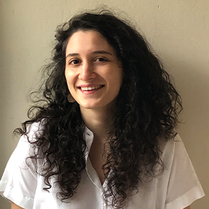 A person with dark curly hair wearing a white shirt standing in front of a cream colored wall.
