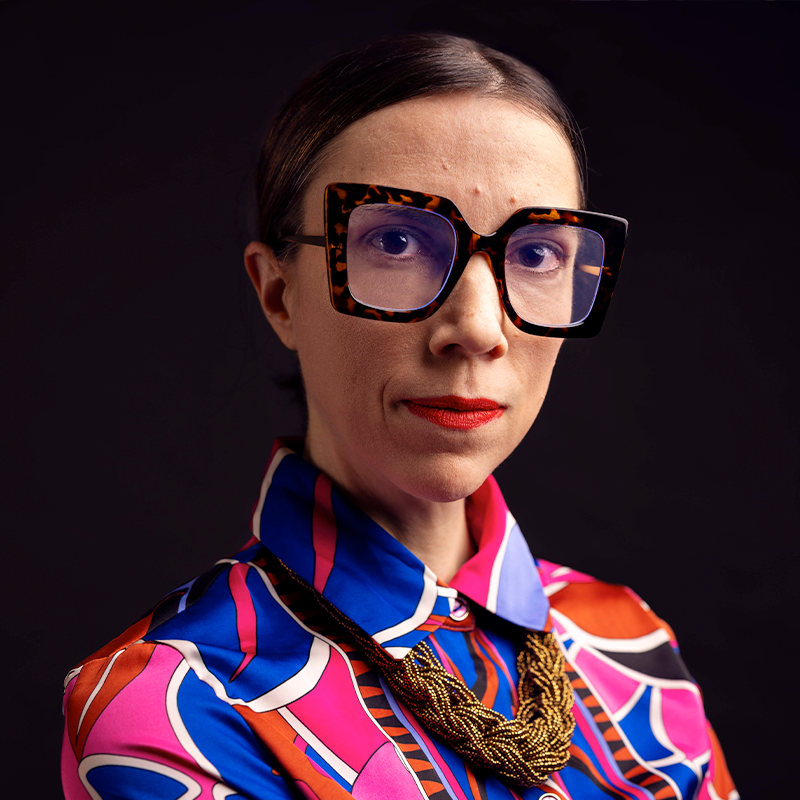 A woman with dark rimmed classes brown hair and a colorful shirt.