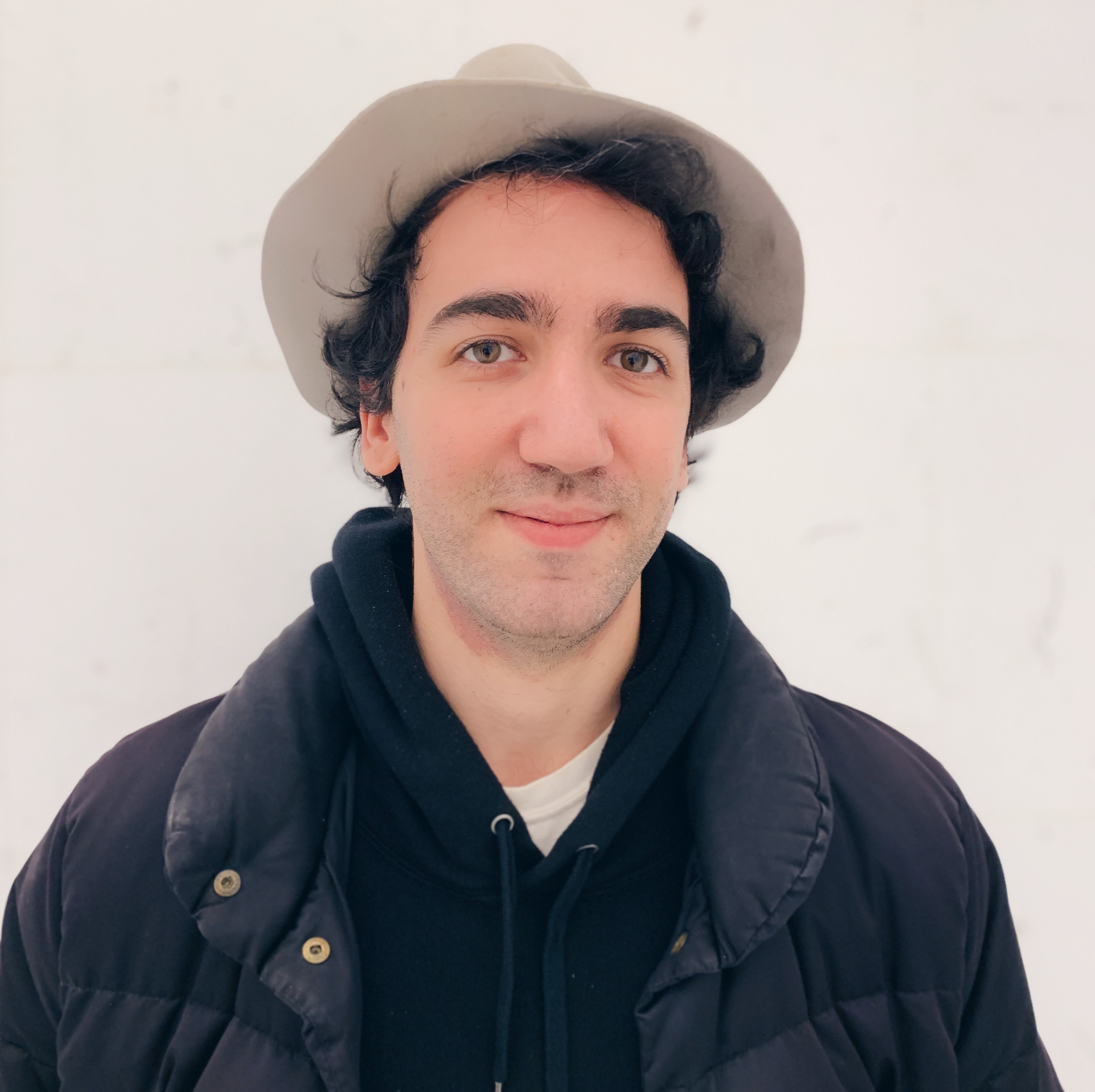 A person with short dark hair wearing a coat and har standing in front of a light background. 