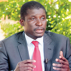 photo of person wearing a black suit, white shirt, and red tie talking with their hands and looking to the right of the camera