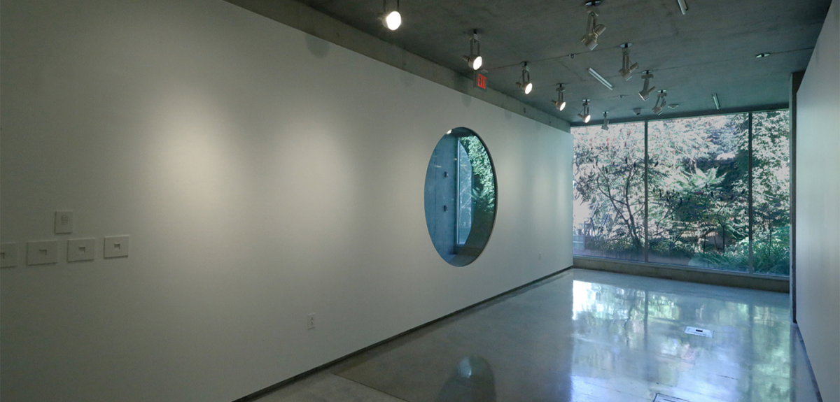 Room with a white wall with floor to ceiling round window and north-side glass wall revealing an outside garden.