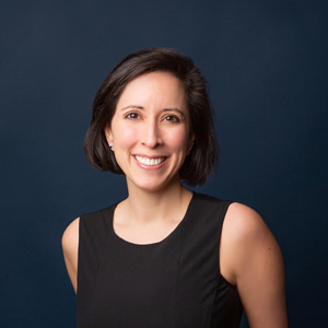 person with black hair in a black shirt with exposed shoulder smiling at the camera against a dark blue background