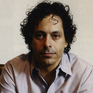 A person wearing a light purple button down shirt in front of a cream colored wall.