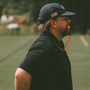 A light skinned man with brown hair and a beard stands in profile on a sports field, wearing a black t-shirt, a blue baseball cap with the American flag on it, and holding an orange whistle in his mouth.