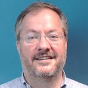 A man with glasses and a light blue shirt in front of a blue background smiles at the camera.