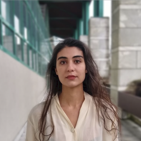 A person with long dark hair standing outside next to a building wearing a white shirt.