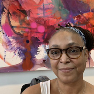 A woman sitting in front of a colorful painting, with dark hair pulled back in a bun, wearing round glasses and a white tank top