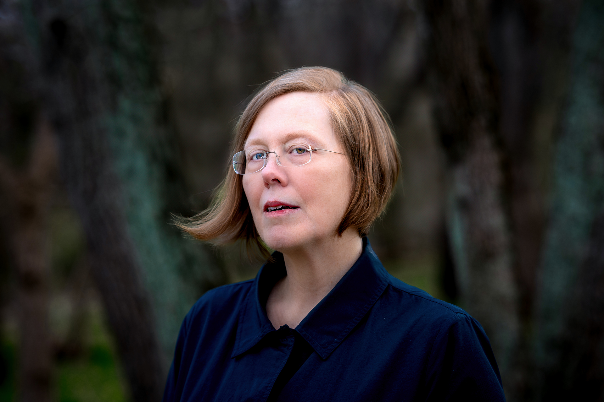 Woman with chin-length strawberry blonde hair wearing a blue jacket in a dark forest
