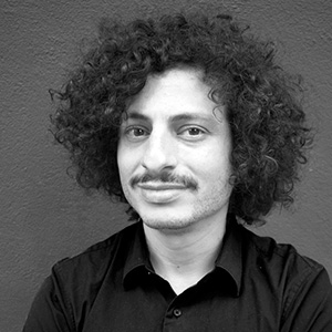 A person with thick curly dark hair and mustache, a dark top