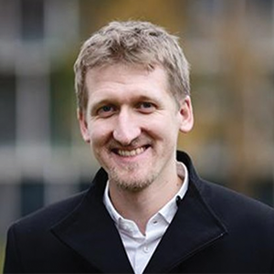 A man in a dark jacket and white shirt smiles at the camera.