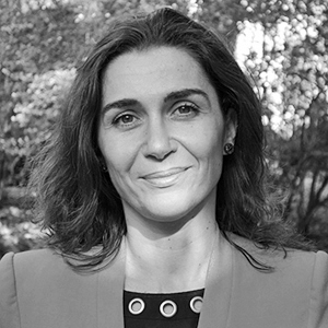 A person with long dark hair wearing a jacket, leafy background.