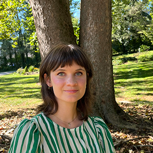 A woman with brown hair and a striped green and white shirt standing in front of a tree looks at the camera.