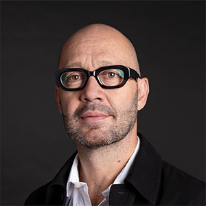 A man with glasses in a black blazer looks at the camera. 