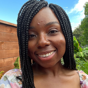 An African American woman with braided hair smiles in front of a grass, brick, and blue sky background.