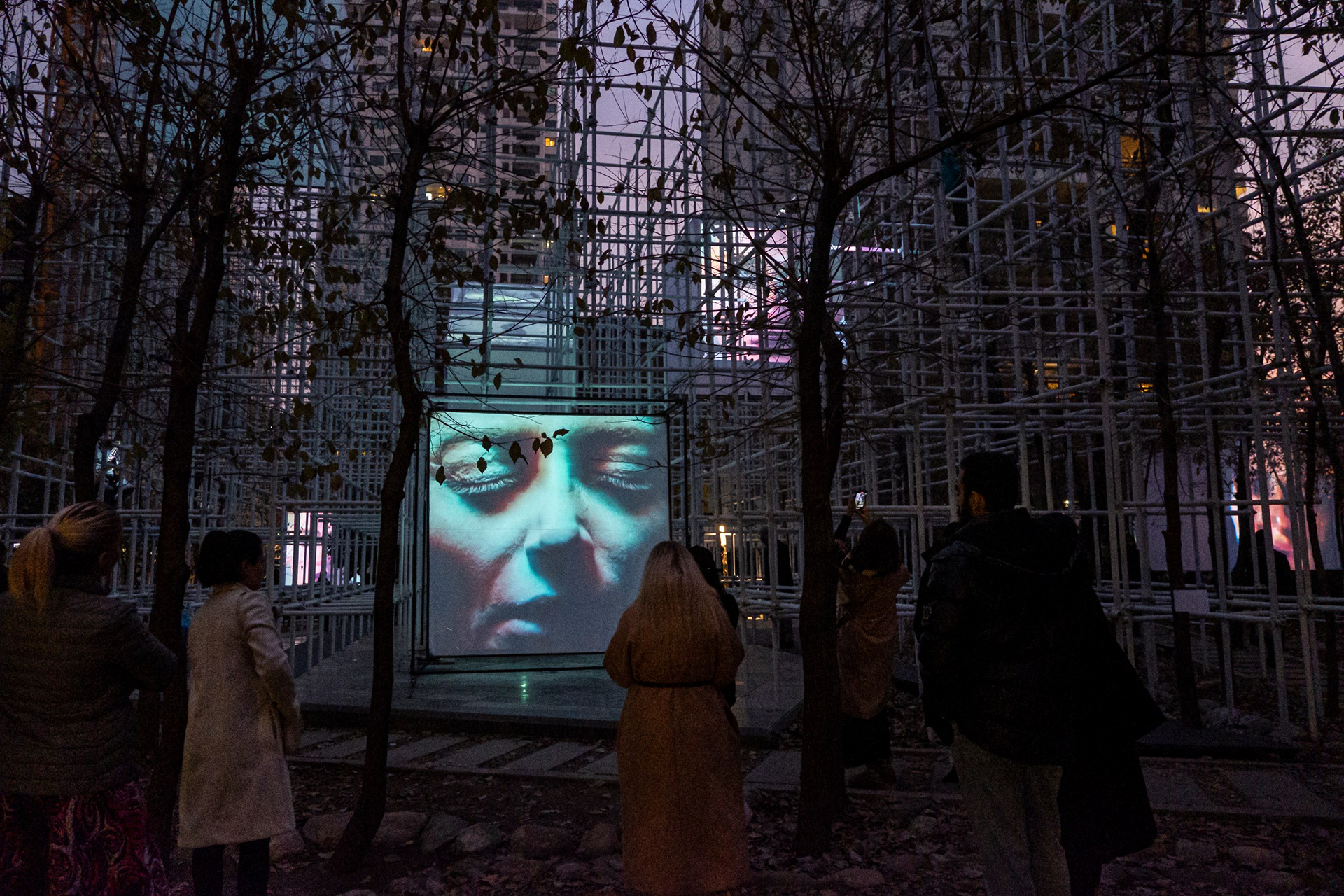  people gathered around an outdoor video projection of a woman's face 