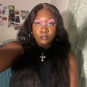 A person with long black hair and multiple facial piercings and bright purple eyeshadow, wearing a black tank top and a cross necklace.