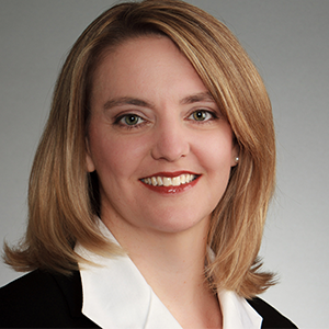 A woman with shoulder-length hair in a black blazer and white shirt smiles at the camera.