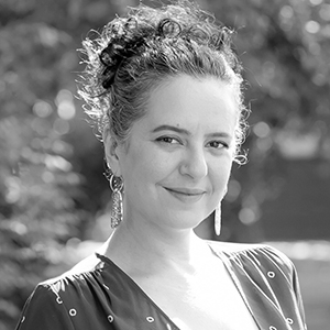 Person smiling, long curly hair in a ponytail, leafy background.