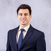 man with dark hair smiling wearing a blue suit and tie