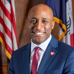 photo of person in blue suit, white shirt, and red tie smiling at the camera with two flags in the background
