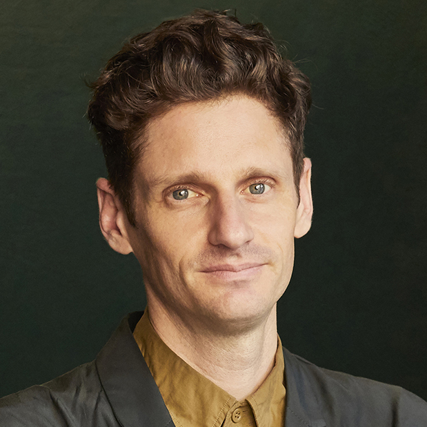 A portrait of a man with short brown hair and blue eyes, wearing a dark green blazer over a mustard-colored shirt. He is smiling slightly and looking directly at the camera, set against a dark green background