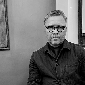 A man with short curly salt-and-pepper hair and light stubble. He has black framed glasses and is wearing a black button down shirt.