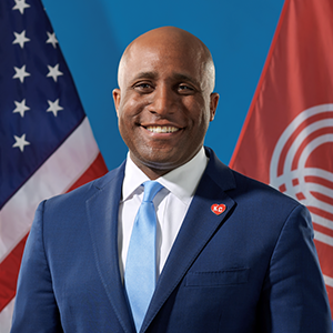 A man in a dark blue suit and light blue tie smiles at the camera.