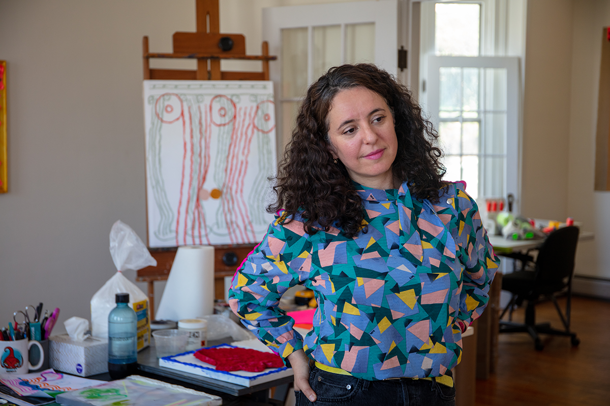 Person with long, dark, curly hair wearing geometrically patterned blouse standing with hands on hips and art supplies in background