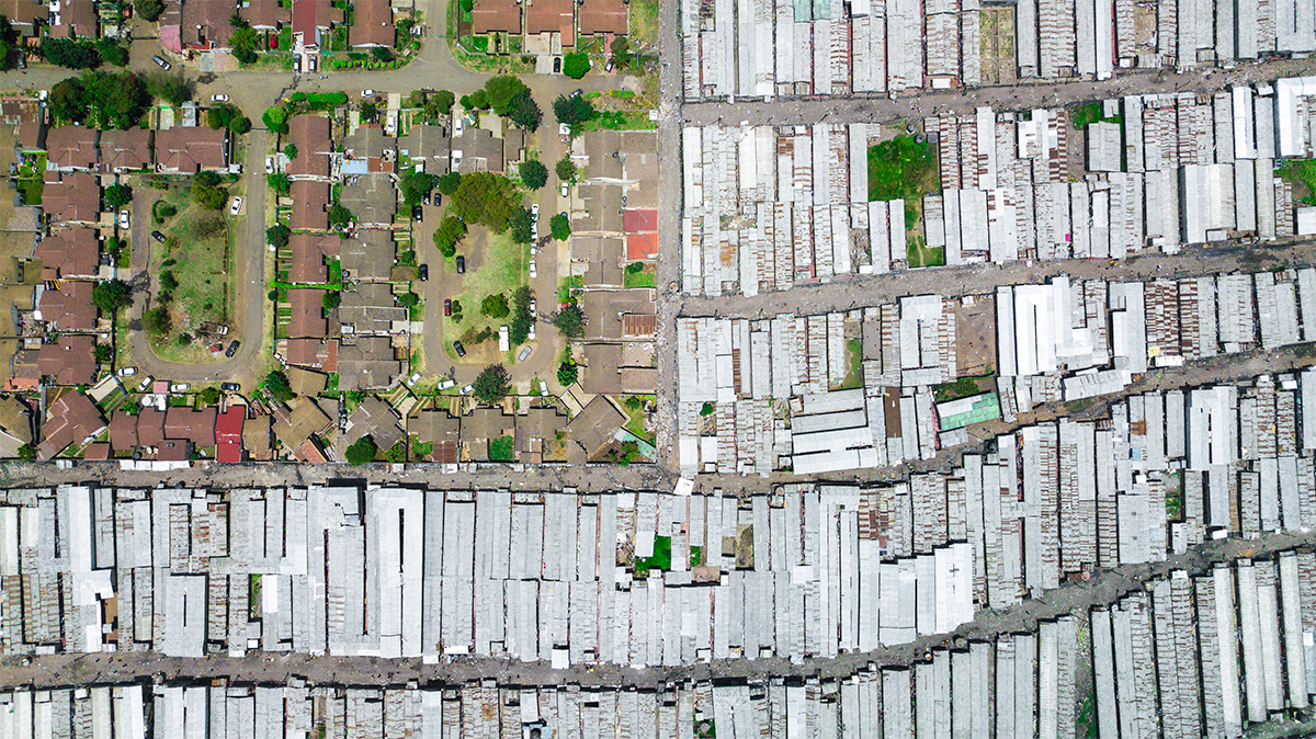 A large array of sheet metal roofed buildings in dirt next to neighborhood of houses with grass and trees