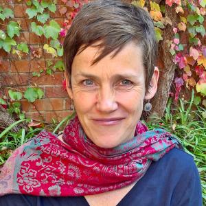 person with short hair and pink scarf looking at the camera against a brick background with leaves