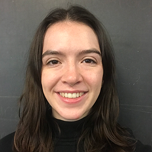 Person with feminine facial features, light skin, and long brown hair wearing black turtleneck smiles wide in front of a black background.
