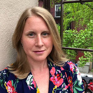 A woman with shoulder length dark blonde hair and blue eyes, wearing a floral patterned shirt and standing in front of a window.