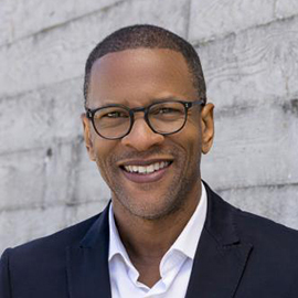 A man wearing black rimmed glasses, a black blazer, and white collared button down shirt. 