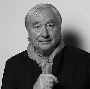 a white man with white hair wearing a dark suit jacket and scarf