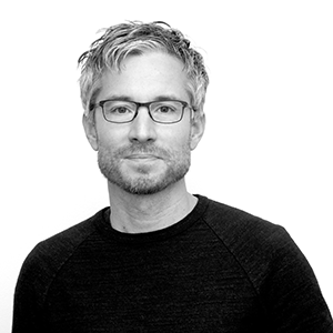 A man with a black shirt and glasses looks at the camera.