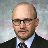 A serious man with a paucity of hair and dark round glasses wearing a suit
