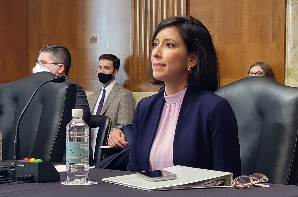 Woman in a blue suit seated at a table in front of a microphone with spectators gathered behind her