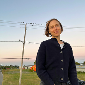 Light skinned person with chin-length blonde hair wearing a navy blue button up shirt smiles against a background of telephone wires, blue and pink sky, and green grass. 