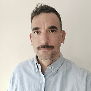 A person with light skin and short brown hair and mustache wearing a light blue collared shirt stands unsmiling in front of a cream background.  