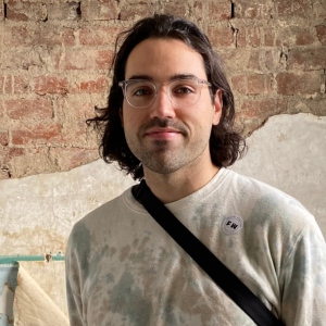 A man wearing glasses looking at the camera, standing in front of a brick wall