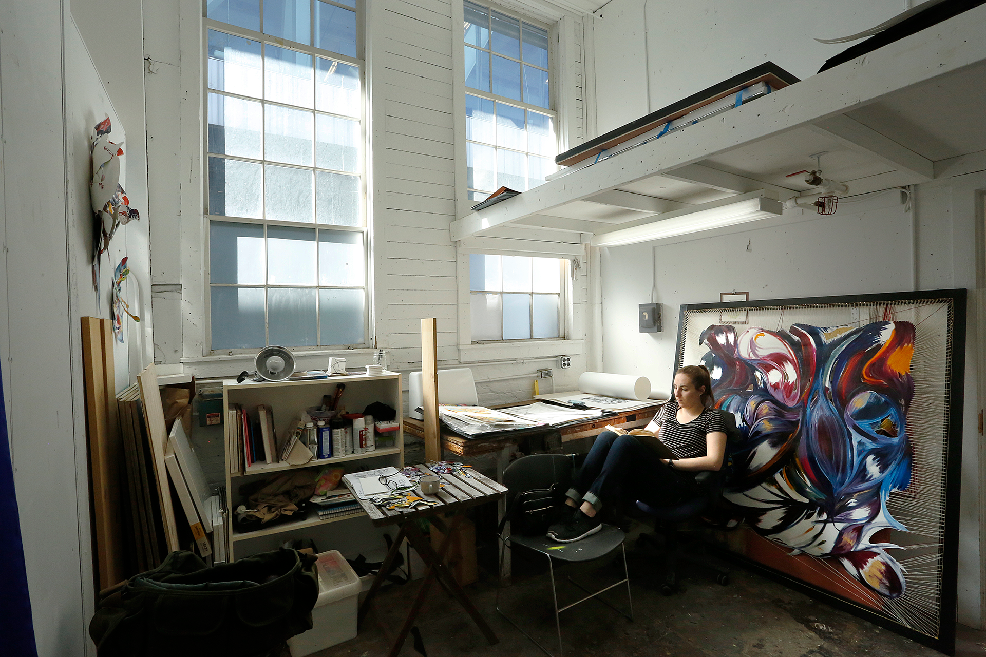 Student curled up in a chair reading a book surrounded by art projects