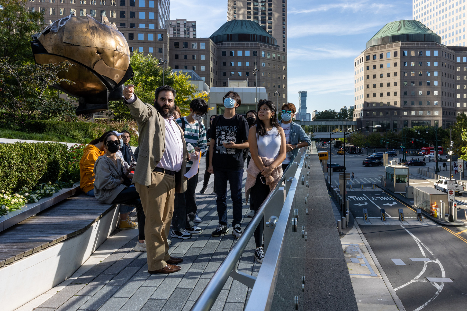 Students gathered on a walkway above an urban street listening to a man in a suit pointing