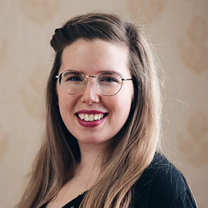 A person with long hair smiling, wearing eyeglasses and a dark top.