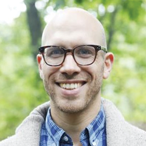 A man with glasses, a white coat and a blue checkered shirt smiles at the camera.