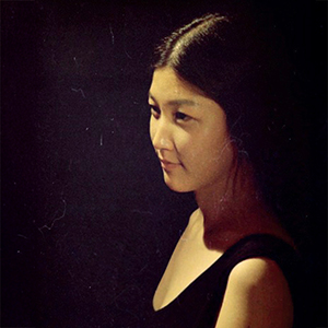 A woman in three-quarter profile, with straight dark hair parted down the middle, and wearing a black tank top.