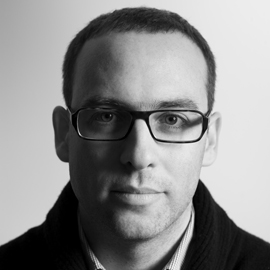 A person wearing glasses and a suit in front of a white wall.